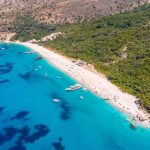 Flygbild över sandstrand med turkost vatten och gröna kullar vid Albaniens kust.