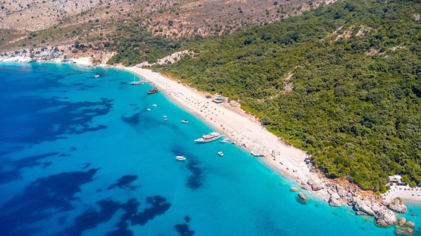 Flygbild över sandstrand med turkost vatten och gröna kullar vid Albaniens kust.