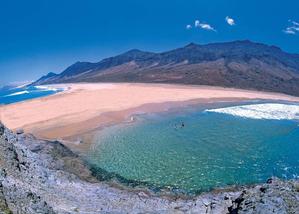 Vidsträckt sandstrand på Fuerteventura med turkost vatten, klippor och karga berg i bakgrunden.