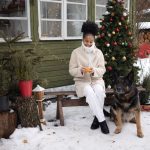 Kvinna och hund sitter utanför ett julpyntat hus i snön.