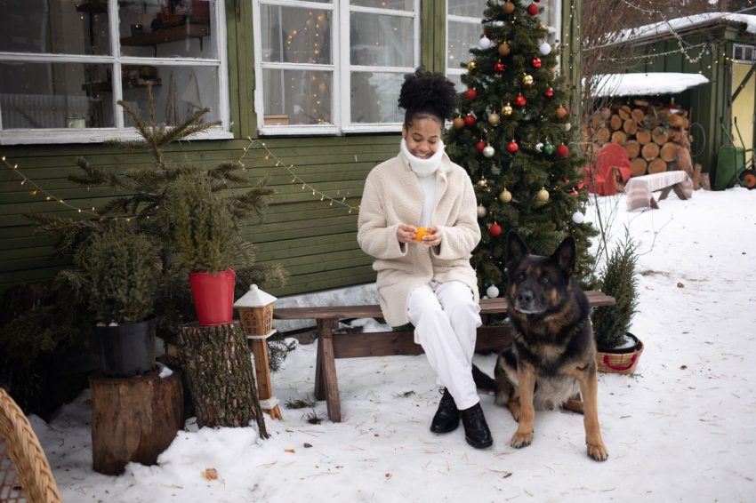 Kvinna och hund sitter utanför ett julpyntat hus i snön.