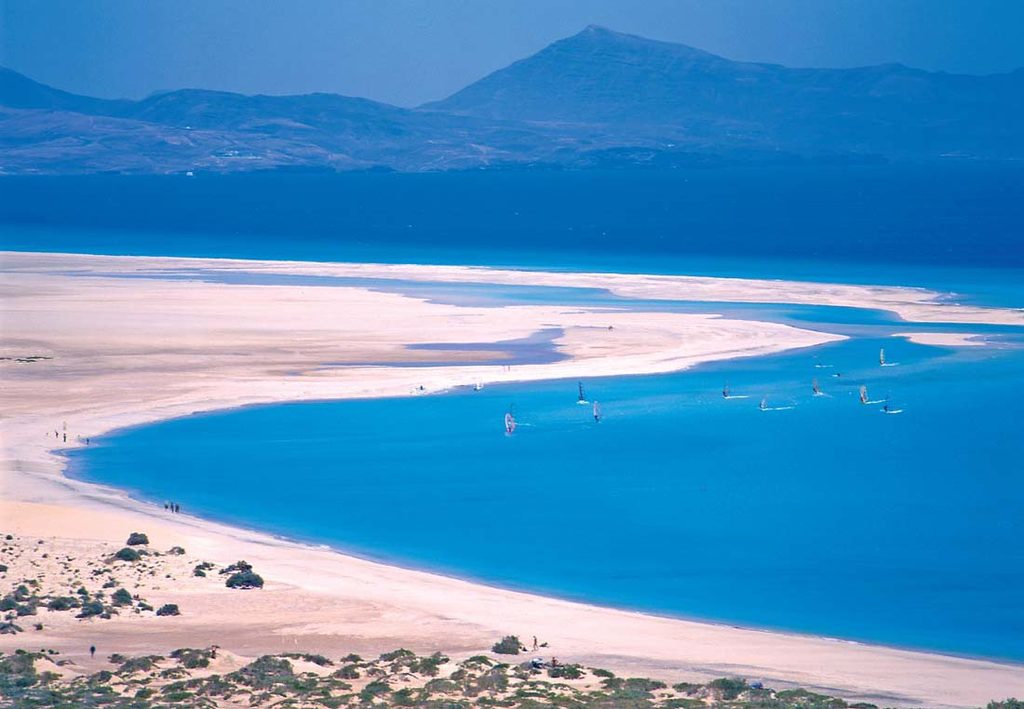 Vy över vita sanddyner och turkost hav på Fuerteventura med vindsurfare i vattnet