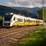 Gult och vitt persontåg på järnväg genom ett grönt bergslandskap i Europa