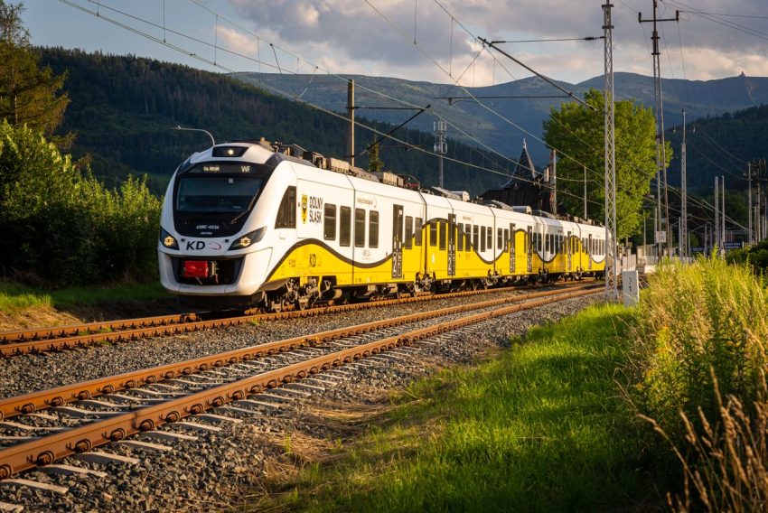 Gult och vitt persontåg på järnväg genom ett grönt bergslandskap i Europa