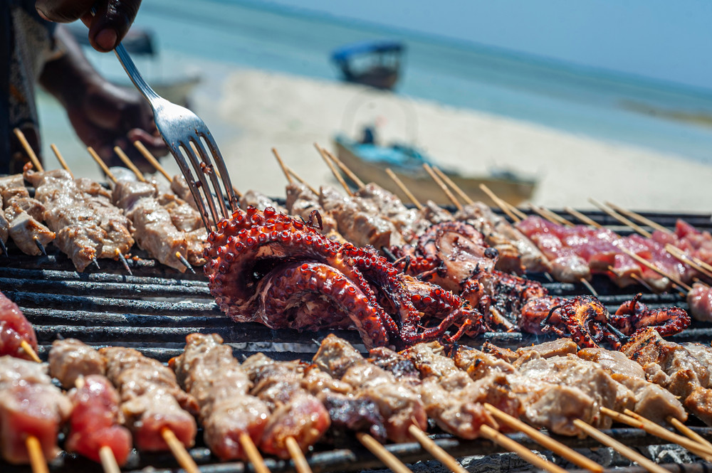 Bläckfisk och grillspett på en grill med stranden och havet i bakgrunden.
