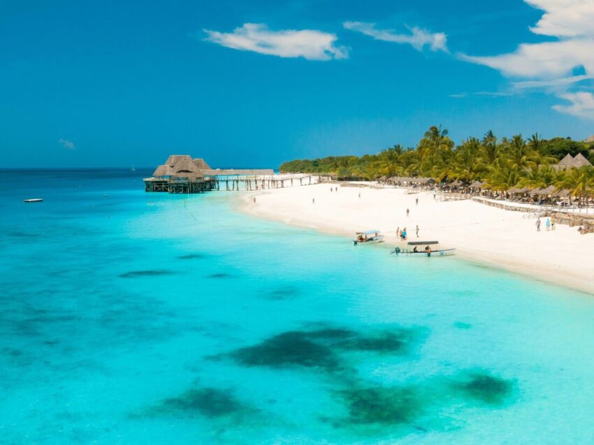 Vit sandstrand med palmer, små båtar och en brygga över turkost vatten på Zanzibar.